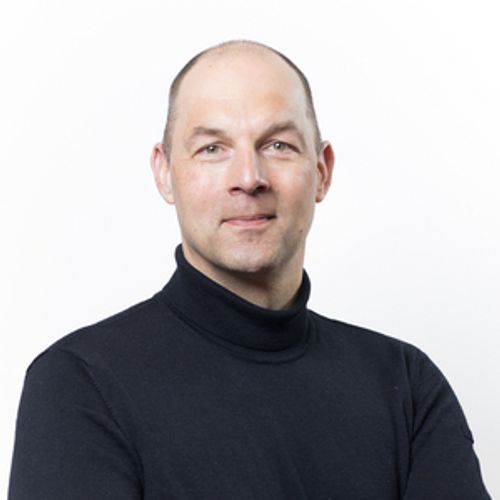 A balding man with green eyes smiles slightly at the camera while wearing a black turtleneck against a plain white background.